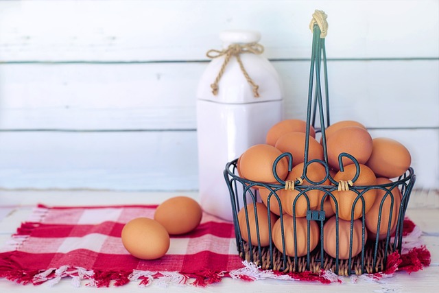 Una cesta de metal llena de huevos morenos sobre una servilleta de cuadros rojos y blancos y una jarra de leche detrás