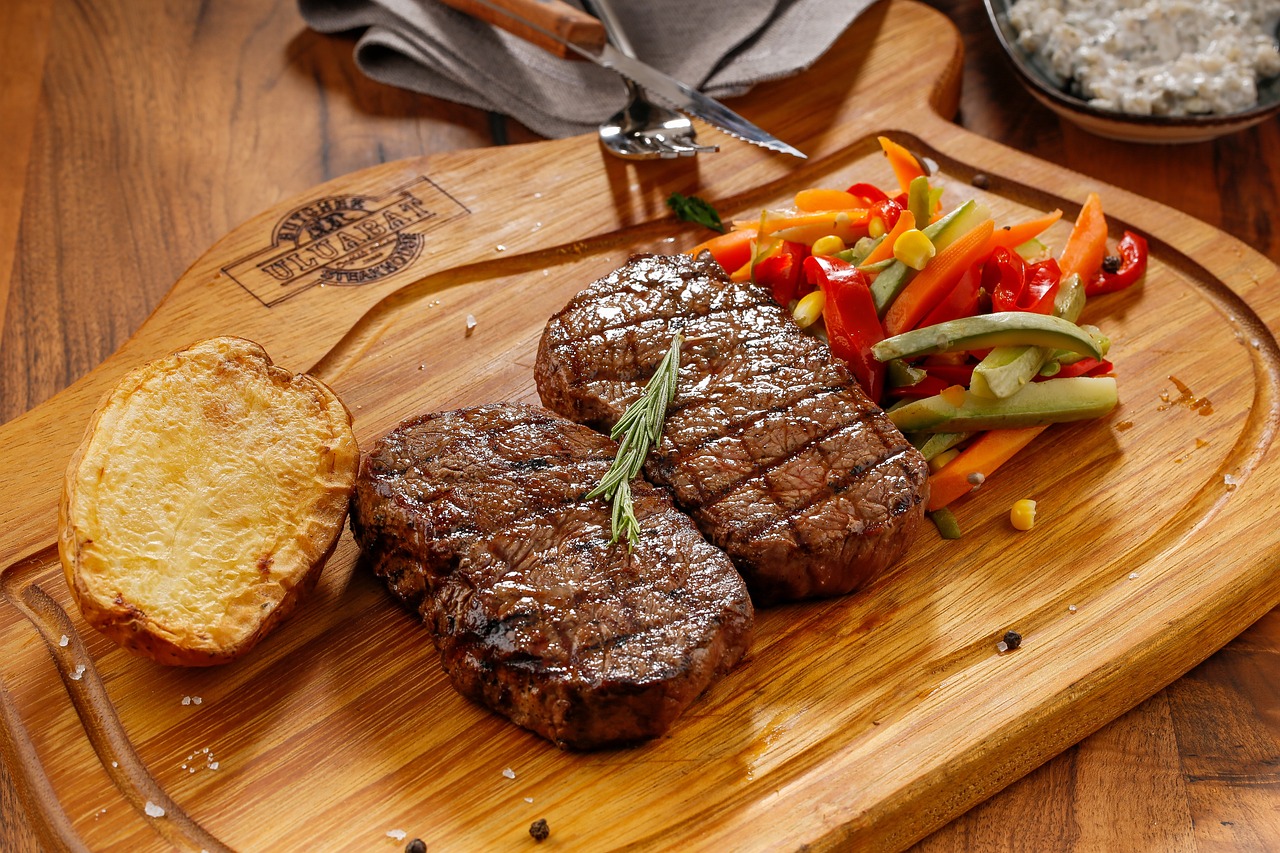 Dos trozos de carne a la parrilla, media patata asada y verduras de guarnición sobre una tabla de madera con un cuchillo y un tenedor